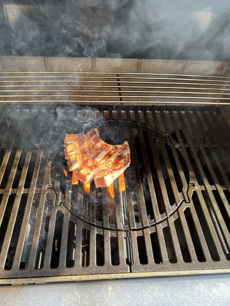 A bone-in pork chop with charred grill marks searing over open flame on a barbecue grill, with smoke rising.